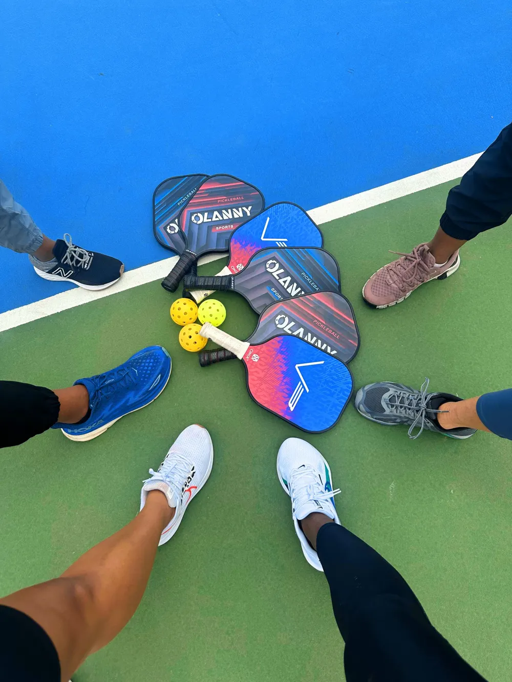 A circle of players' feet and paddles gathered on a green and blue pickleball court